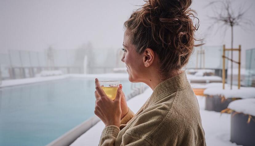 Woman in bathrobe drinks tea by the snow-covered infinity pool.