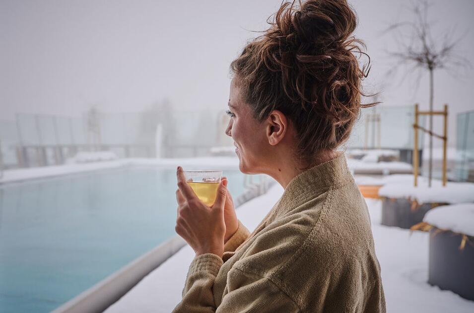 Woman in bathrobe drinks tea by the snow-covered infinity pool.