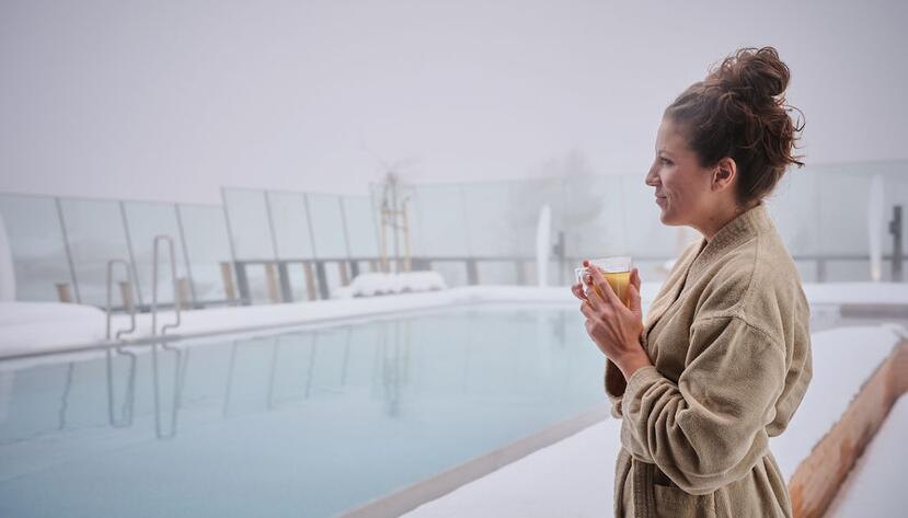 Woman in bathrobe drinks tea by the snow-covered infinity pool.
